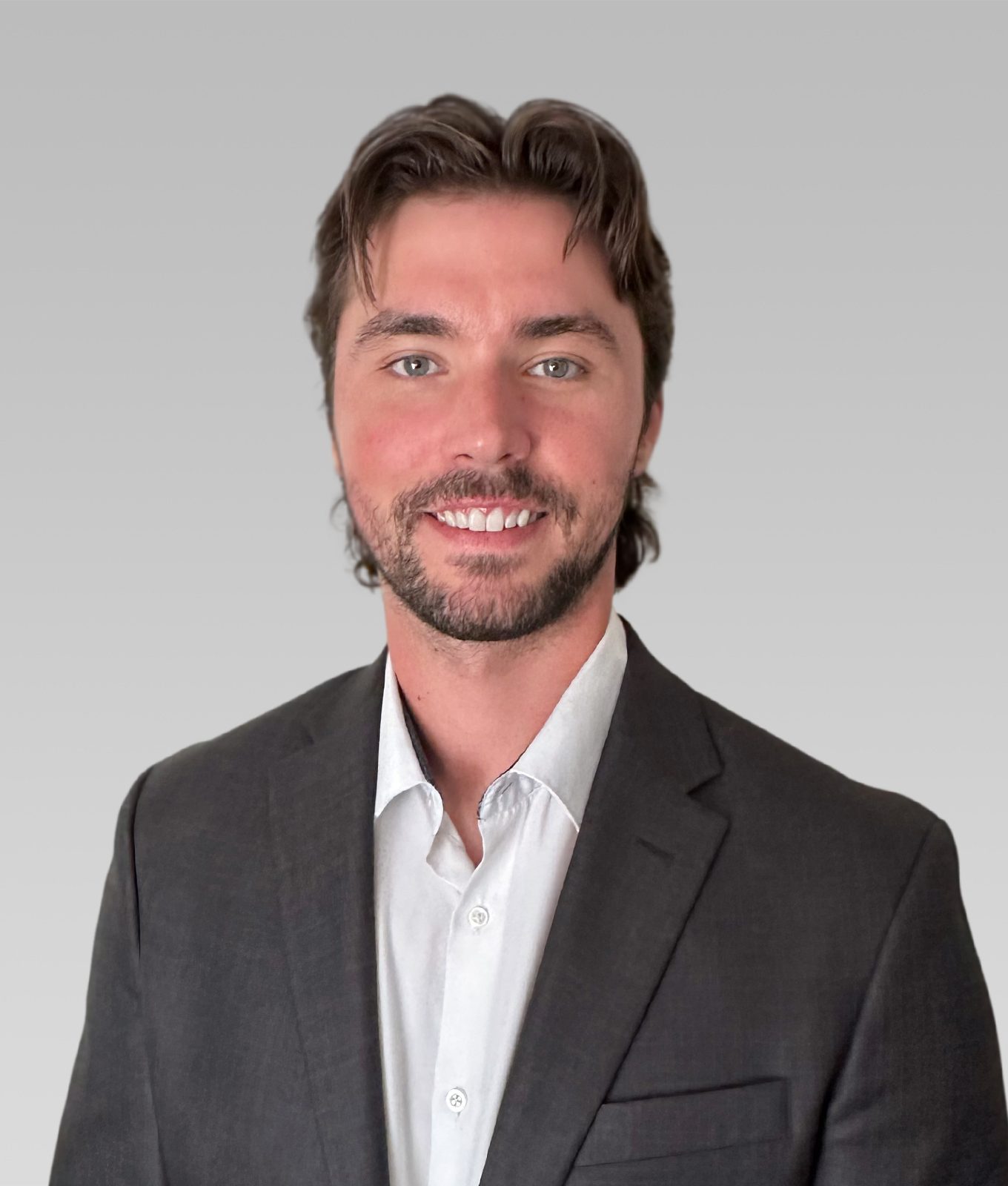 Smiling man with brown hair and beard in a dark suit and white shirt against a gray gradient background.