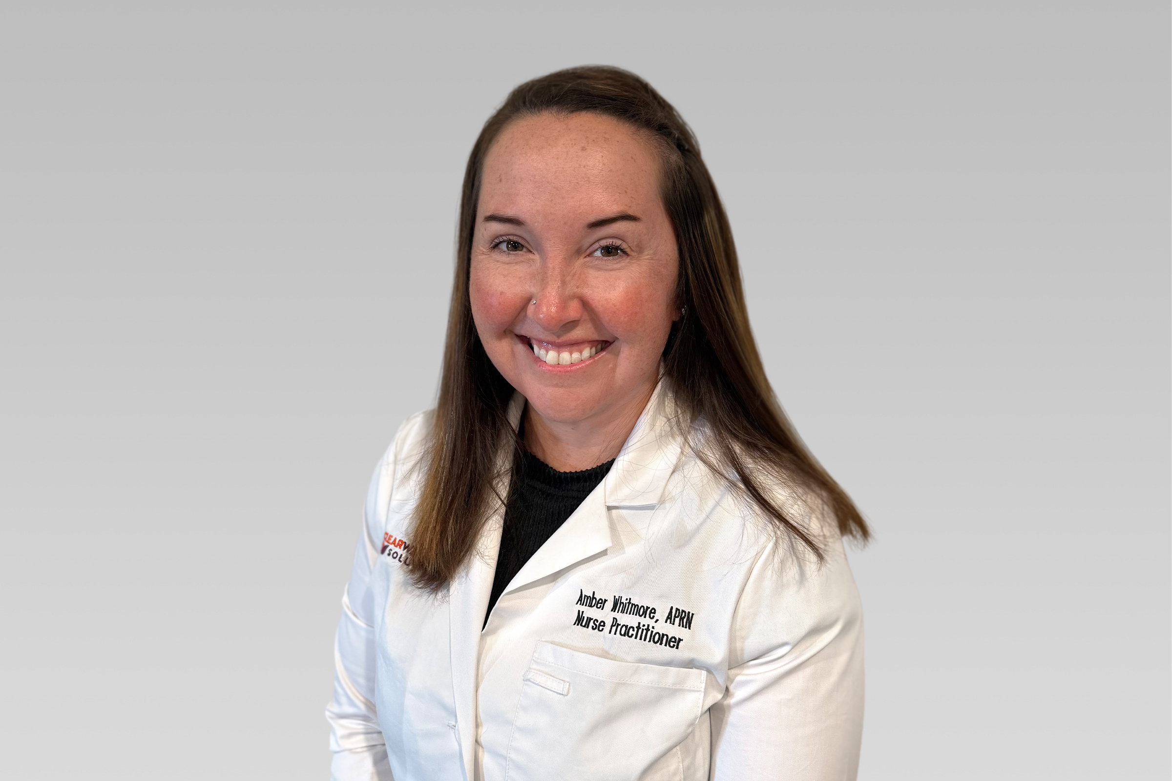 Portrait of Amber Whitmore, APRN, a nurse practitioner, smiling in a white coat against a gray background.