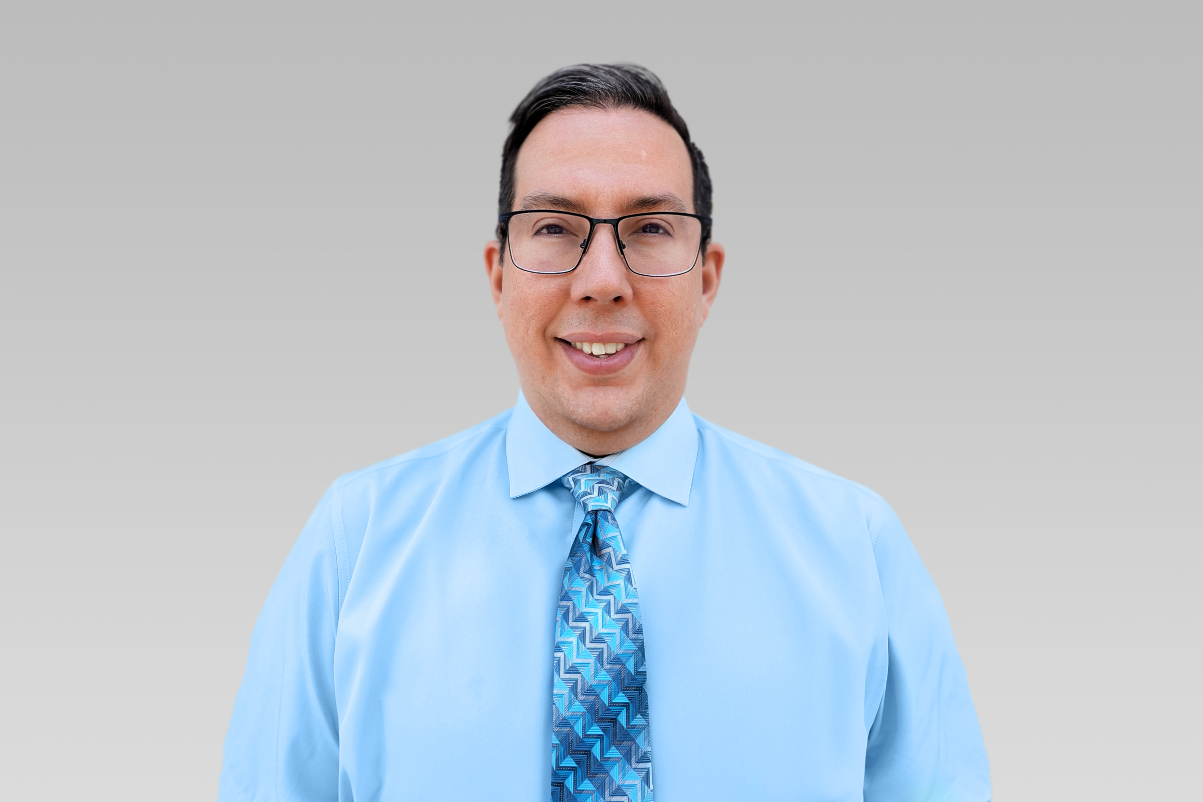 Smiling man in a light blue shirt and patterned blue tie, wearing glasses, against a gray gradient background.