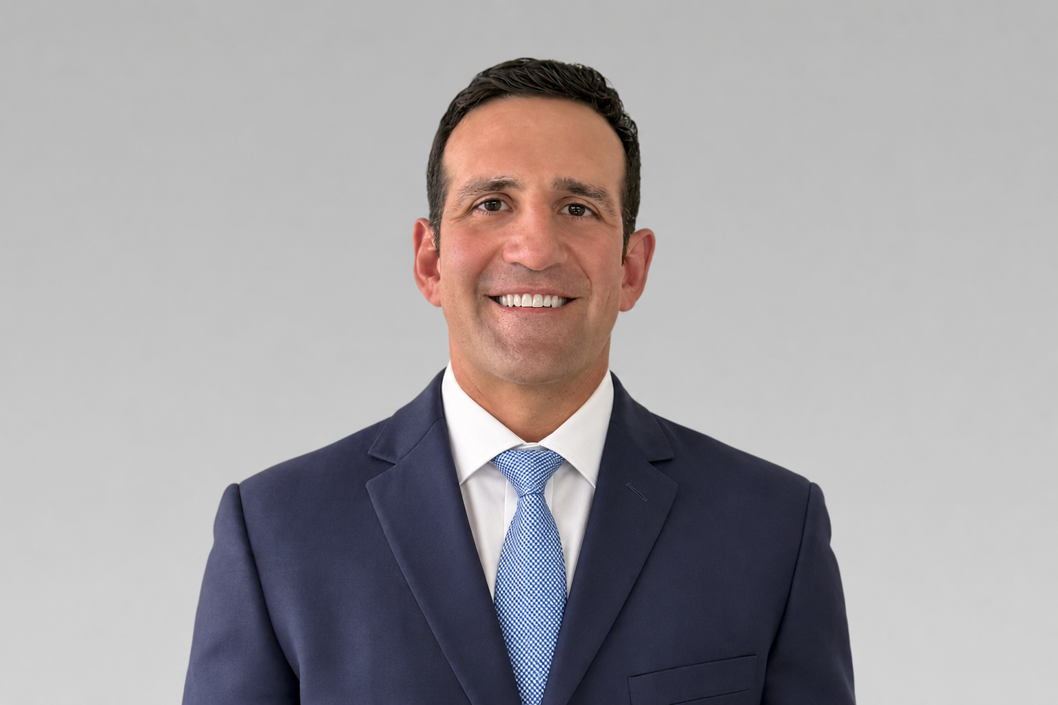 Professional headshot: smiling man in a navy suit, white shirt, and blue tie against a neutral gray background.